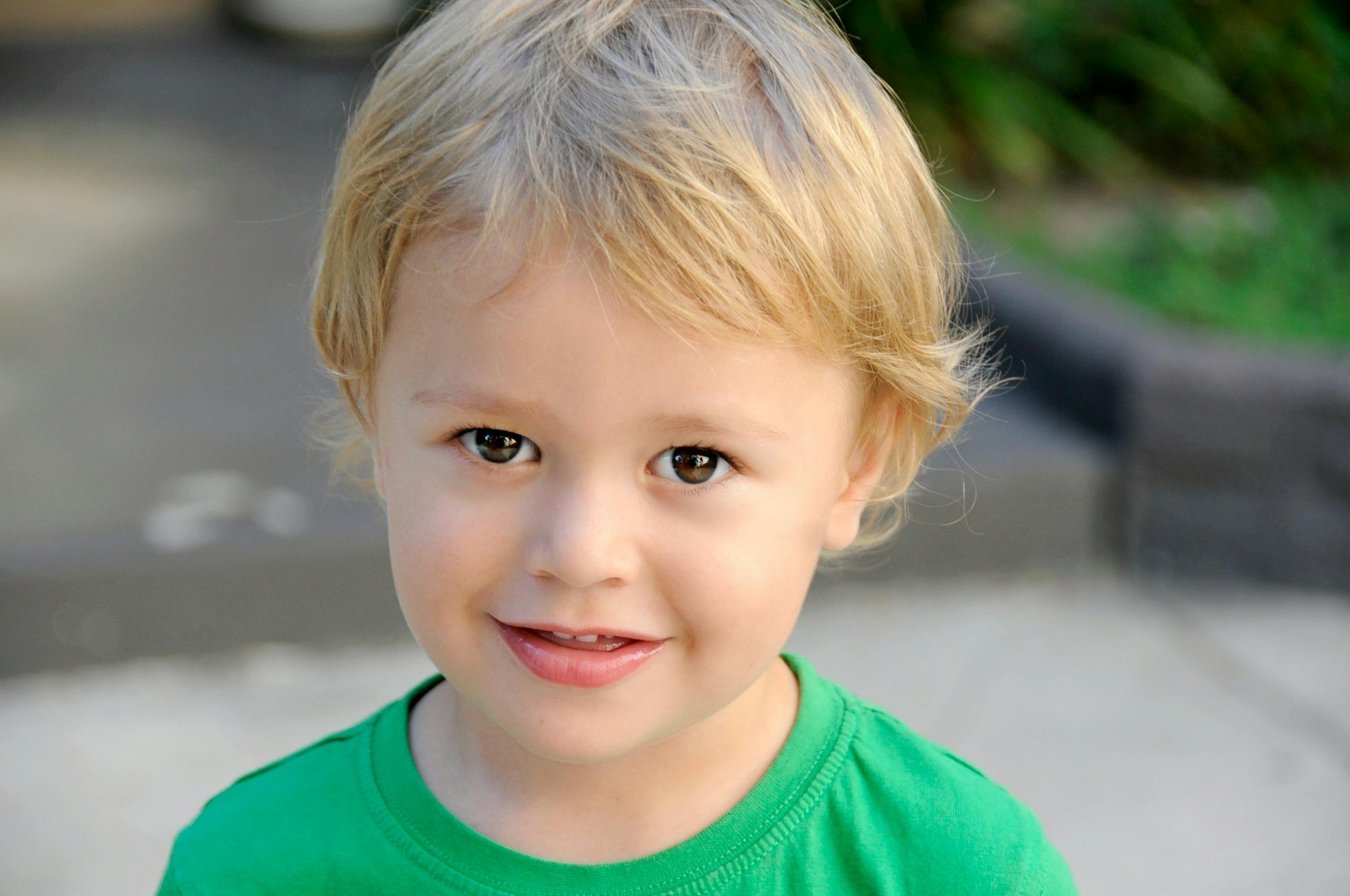 A cheerful blond child smiles outdoors in a green shirt, evoking innocence and joy.